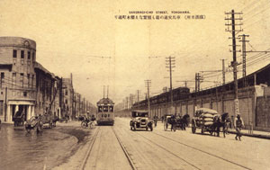 震災復興期の桜木町通り　絵葉書　当館所蔵　左側から荷車、牛車、自転車、市電、自動車、人力車、荷馬車、  電車が確認できる。横浜の街を様々な乗り物が駆けていた。